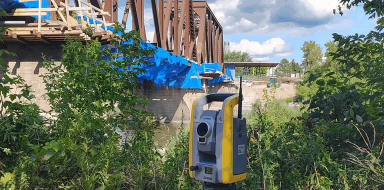 Robotic total station set up in the grass beside a steel truss bridge under construction